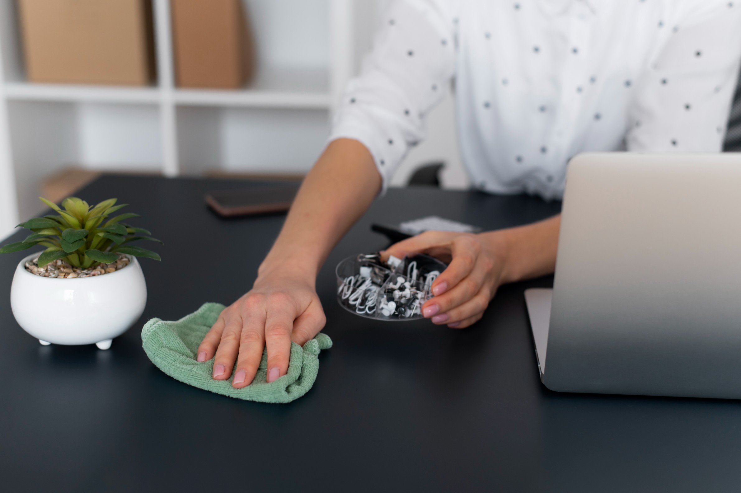 Person methodically cleaning a desk with a cloth beside a laptop and plant, representing the ‘chunk by chunk, top to bottom’ cleaning method for achieving order, focus, and calm through structured cleaning.